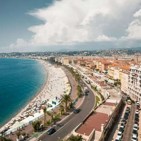 Vue sur la Promenade des Anglais à Nice, entre mer azur, plage animée et toits ocres, sous un ciel nuageux et lumineux.