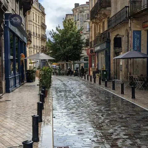 Rue pavée mouillée après la pluie, bordée de cafés et boutiques, avec une ambiance paisible et reflets lumineux sur le sol.