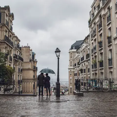 Un couple sous un parapluie contemple Paris depuis une rue pavée bordée d’immeubles haussmanniens, par un jour de pluie.