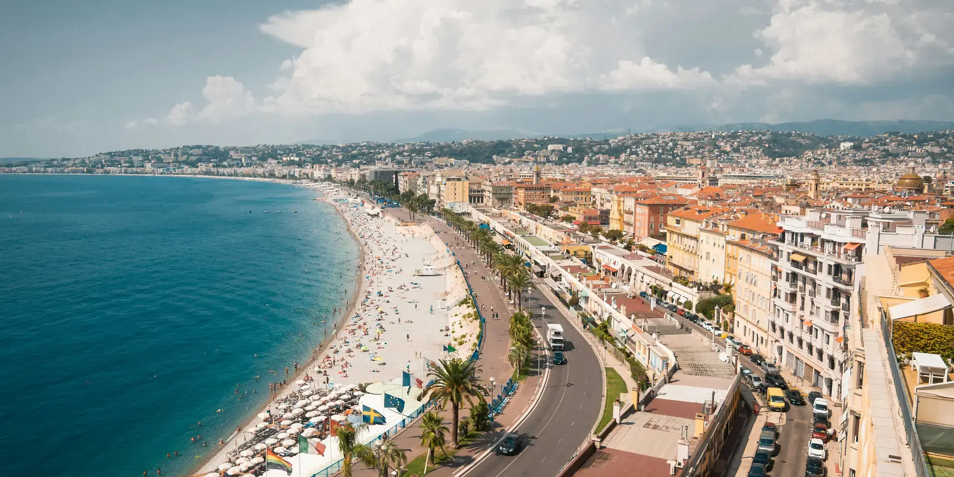 Vue sur la Promenade des Anglais à Nice, entre mer azur, plage animée et toits ocres, sous un ciel nuageux et lumineux.