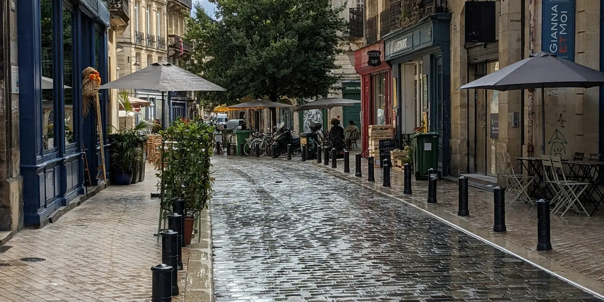 Rue pavée mouillée après la pluie, bordée de cafés et boutiques, avec une ambiance paisible et reflets lumineux sur le sol.