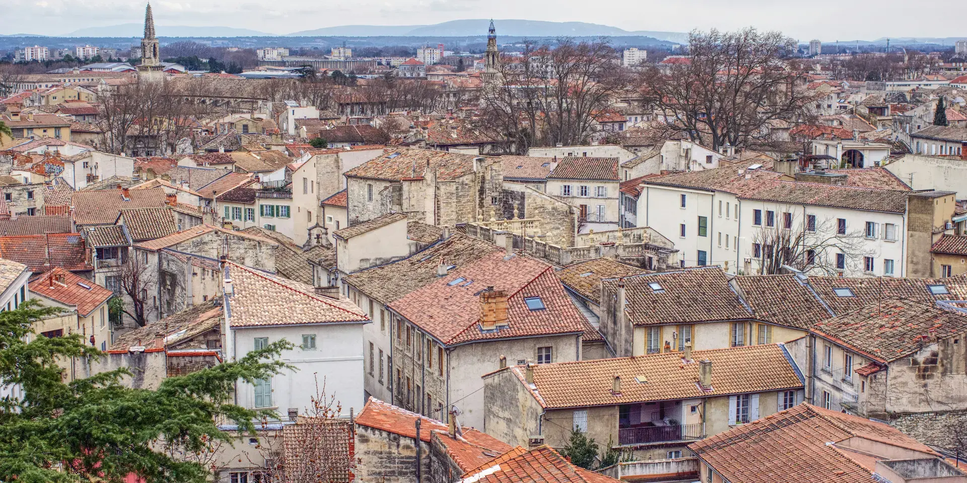 Vue sur les toits en tuiles d’une ville du sud de la France, avec ruelles et clochers, sous un ciel nuageux.