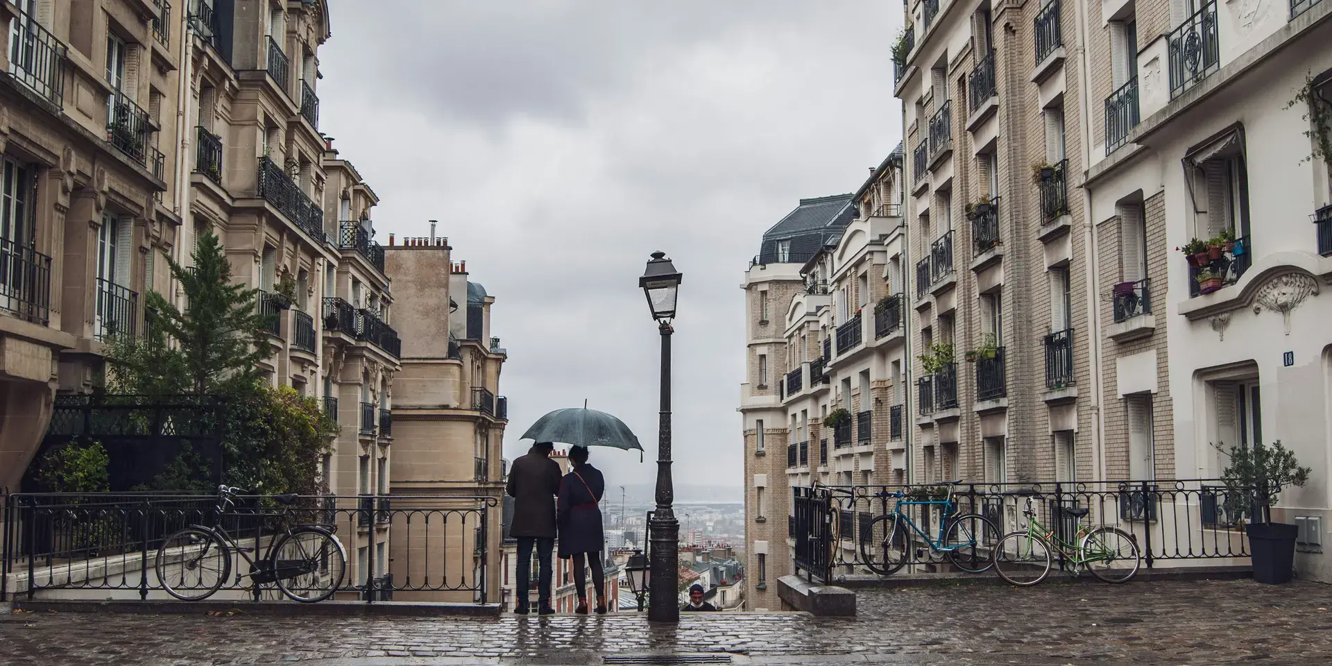 Un couple sous un parapluie contemple Paris depuis une rue pavée bordée d’immeubles haussmanniens, par un jour de pluie.