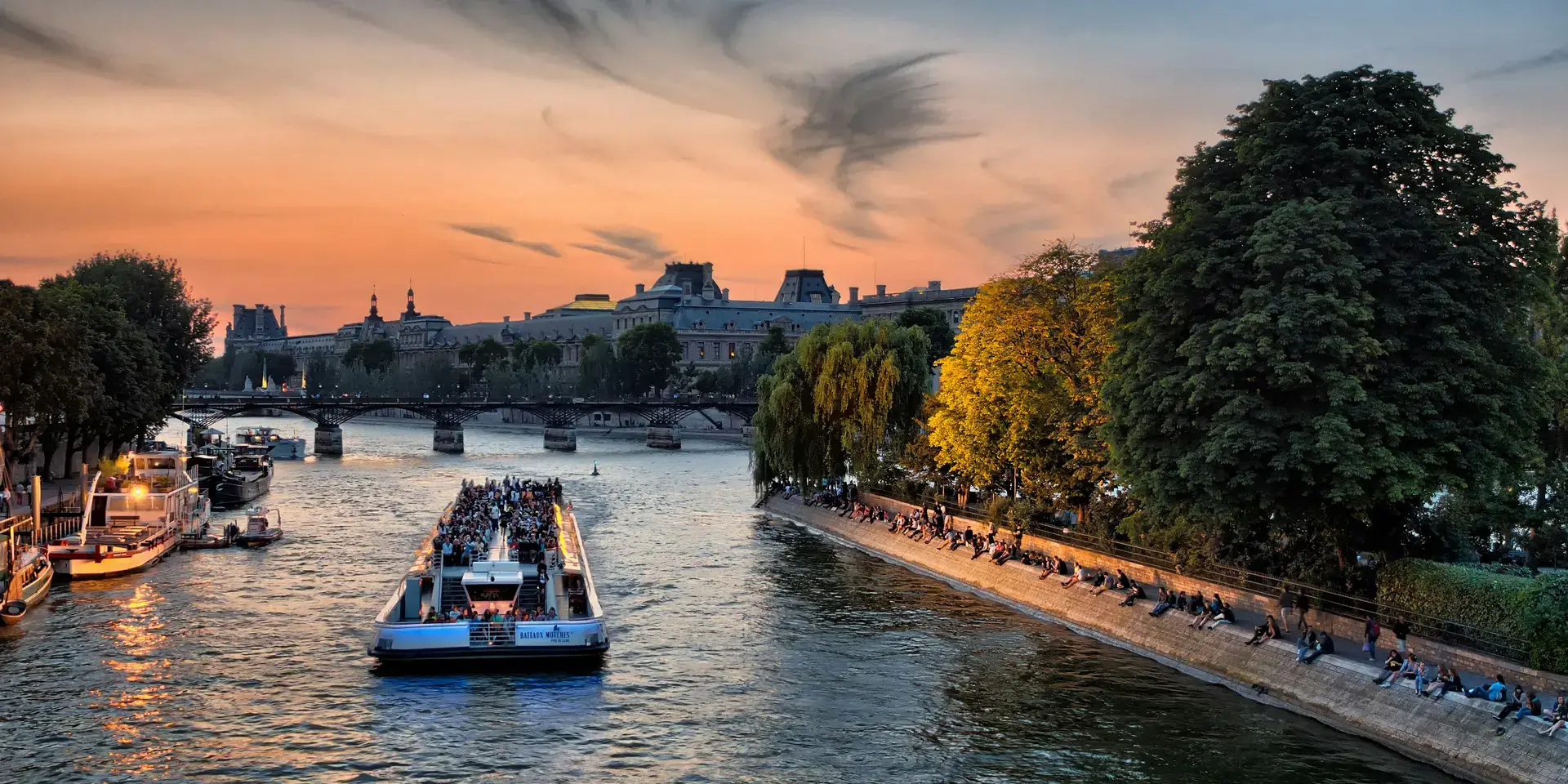 Croisière sur la Seine au coucher du soleil, avec vue sur les quais animés et les monuments parisiens sous un ciel doré.